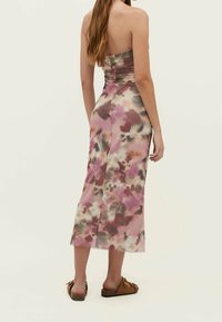 Nainen, jolla on pitkät hiukset, pukeutunut olkaimettomaan vaaleanpunaisiin ja ruskeisiin batikkikuosisisiin midi-mekkoon ja ruskeisiin liukuvaatteisiin, seisoo yksinkertaista taustaa vasten.