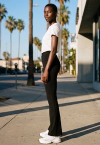 Høj person står sidelengs på en solbeskinnet fortov iført hvid skjorte, sorte bukser og hvide sneakers med palmer og bygninger i baggrunden.