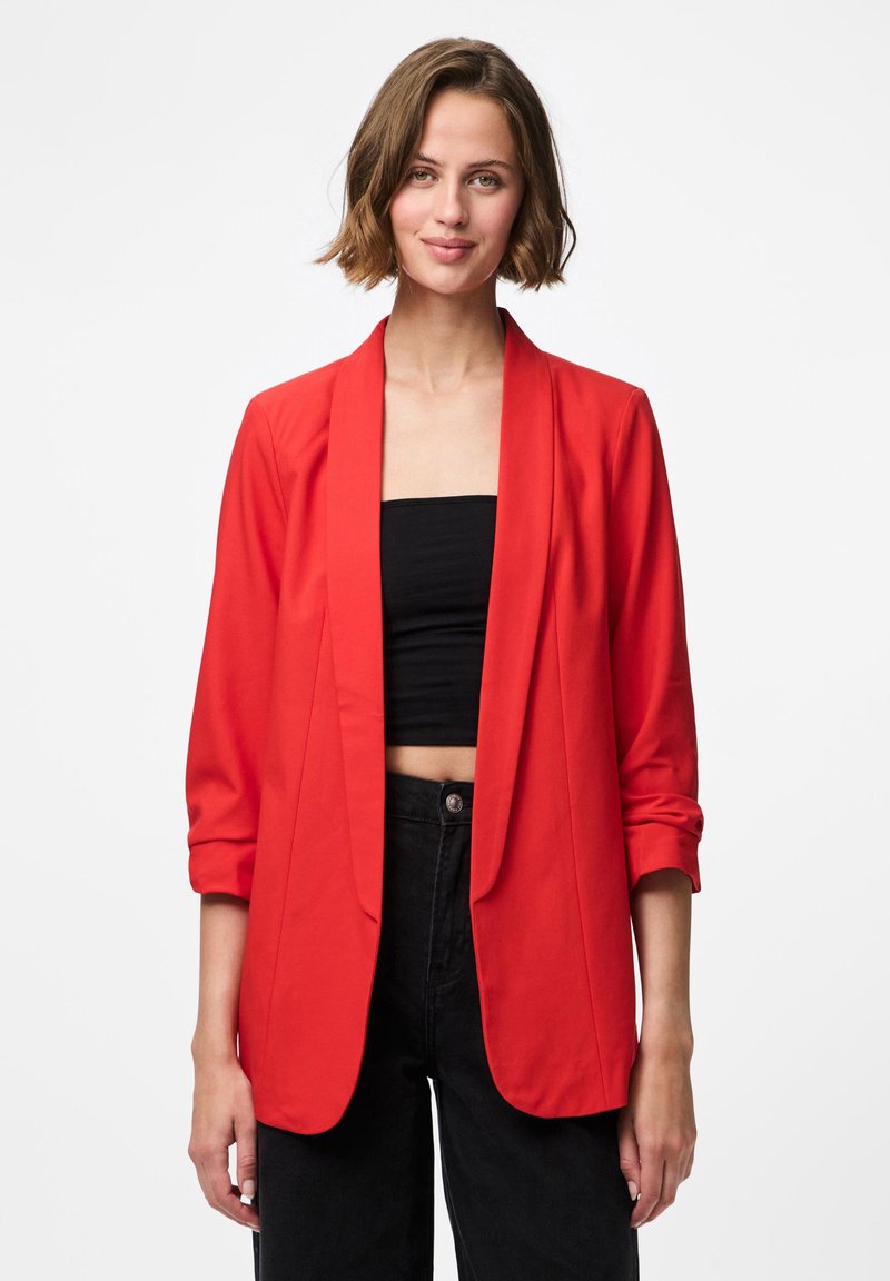 Červený blazer s šálovým límcem, dlouhými rukávy ohrnutými k loktům a otevřeným předním dílem. Nosený přes černý cropped top a černé kalhoty s vysokým pasem.