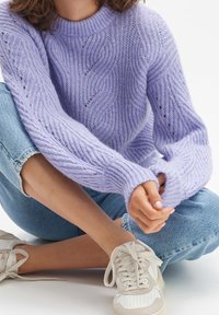 Kvinna som bär en lavendelfärgad kabelstickad tröja, ljusblå jeans och vita sneakers, sittande med korslagda ben på ett vitt golv.