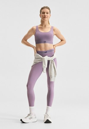 Nainen seisoo itsevarmasti, päällään violetti urheilurintaliivit ja leggingsit, valkoiset lenkkarit sekä vyötärön ympärille sidottu vaaleanharmaa collegepaita.