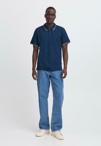 V visokem moškem, ki nosi mornarsko modro polo majico, svetlo modre kavbojke in bele superge, ki stoji ob enobarvni svetli ozadju.