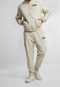 Světlé béžové teplákové soupravy se zipovou bundou a kalhotami s elastickým pasem, vyrobené z texturované látky s logem na hrudi a stehně.