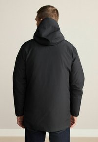 Sort vandtæt jakke med hætte, præsenterer et elegant design og en glat tekstur. Jakken går ned til lårene og har lange ærmer.