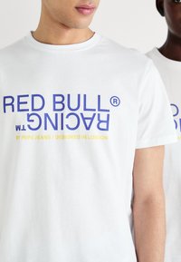 Balta marškinėlių su trumpomis rankovėmis, ant kurių ryškiai mėlynu šriftu yra užrašas „RED BULL RACING“, su maža geltona dizaino detale. Pagaminta iš minkštos medvilnės, laisvą siluetą.