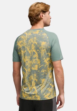 Uomo con capelli biondi corti che indossa una camicia a maniche corte con motivo botanico verde e giallo, di spalle alla fotocamera.