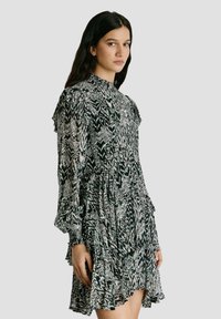 Ženska z dolgimi temnimi lasmi, oblečena v črno-belo obleko z vzorcem in dolgimi rokavi z volančki, stoji pred enostavnim sivim ozadjem.