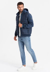 Mörkblå vadderad jacka med huva, med dragkedja och sidofickor. Matchad med ljusblå slitna jeans och vita sneakers.