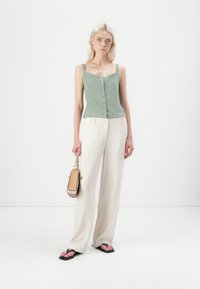 Top a canotta aderente verde chiaro con bottoni, abbinato a pantaloni a gamba larga color crema. La modella tiene in mano una piccola borsa beige e indossa ciabattine nere.