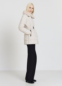 Cappotto imbottito beige con colletto in pelliccia sintetica, vita con cintura e tasche con zip, abbinato a pantaloni neri a zampa d'elefante e tacchi neri.