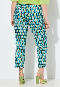 Persona vistiendo pantalones con estampado geométrico vibrante en azul, verde y blanco, combinados con sandalias de tacón verde, vista desde atrás.