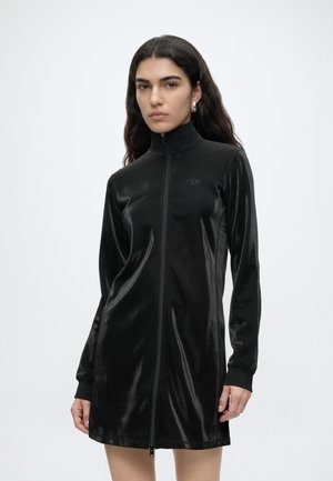 Femme portant une robe noire en velours à manches longues avec fermeture éclair, col montant et boucles d’oreilles argentées en forme d’anneaux, debout devant un fond uni.