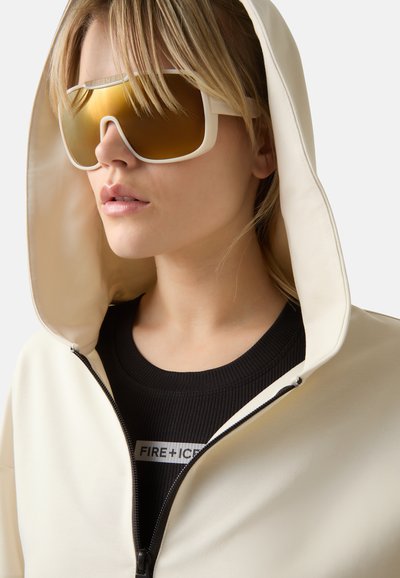 Sonnenbrille mit goldenen Spiegellinsen und einem weißen, breiten Rahmen, kombiniert mit einem cremefarbenen Zip-up-Hoodie und einem schwarzen gerippten Oberteil mit einem Logo-Tag.