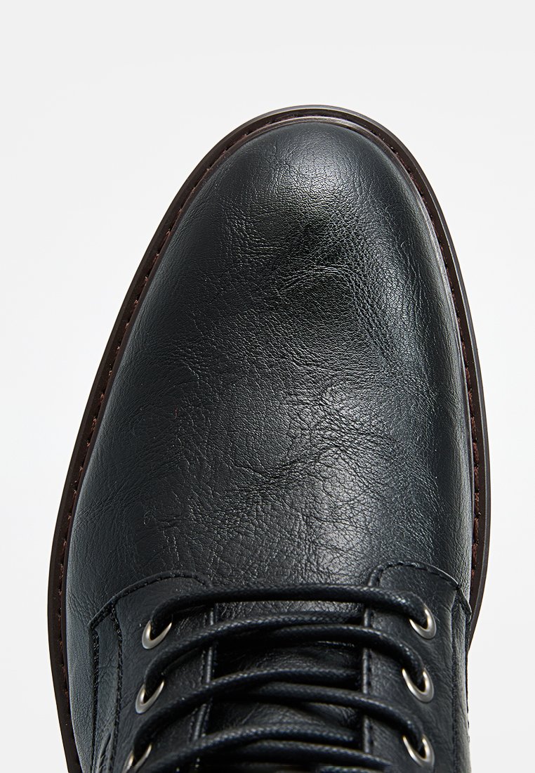 Zapato de cuero negro con puntera redonda, acabado texturizado y ojales plateados para los cordones. Suela de goma con costuras marrones a lo largo del borde.