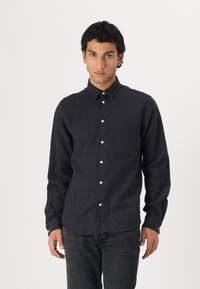 Cămașă lungă de culoare bleumarin închis, dintr-un material texturat, cu guler tip button-down, nasturi albi și un buzunar pe piept. Purtați-o cu blugi gri.