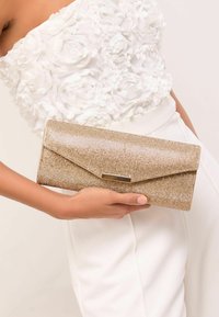Geantă clutch cu sclipici auriu, cu o clapetă triunghiulară și un detaliu accent auriu. Textură metalică netedă, formă dreptunghiulară, ținută într-o mână pe un fundal alb.