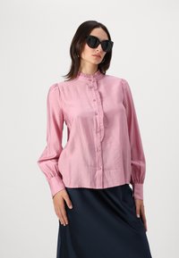 Rosa satengbluse med høy krage, volanger og lange ermer; har knappestolpe og subtil glans. Modellen har på seg svarte solbriller.