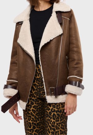 Mujer con chaqueta marrón de piel con forro de piel crema, correas oscuras con hebilla en mangas y cintura, combinada con pantalones de estampado de leopardo y top negro.