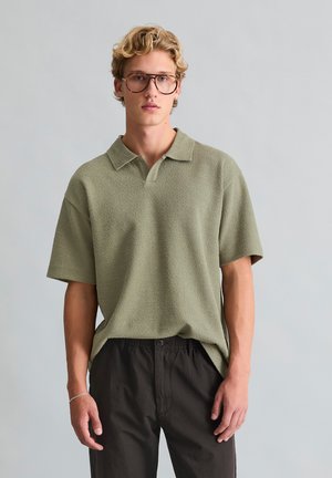 Jonge man met golvend blond haar, die een oversized bruine bril, een olijfgroene gestructureerde polo, een zwarte broek en een zilveren armband draagt, staat neutraal.