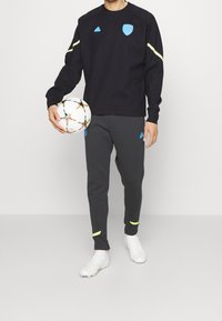 Felpa sportiva nera con dettagli gialli e un'emblema blu, abbinata a pantaloni della tuta grigi con dettagli blu e polsini gialli, mentre tiene un pallone da calcio.