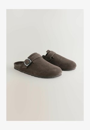Bruine suède klompen met een ronde neus, een brede band en een zilveren gespdetail, voorzien van een gestructureerde rubberen zool.