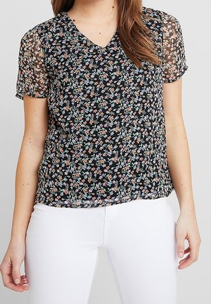 Blouse noire transparente à manches courtes avec petit imprimé floral multicolore, portée avec un pantalon blanc, vue du cou jusqu'aux hanches.
