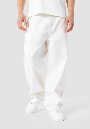 Person iført løse hvide bukser og hvide sneakers, stående med hænderne afslappet langs siden mod en lysegrå baggrund.