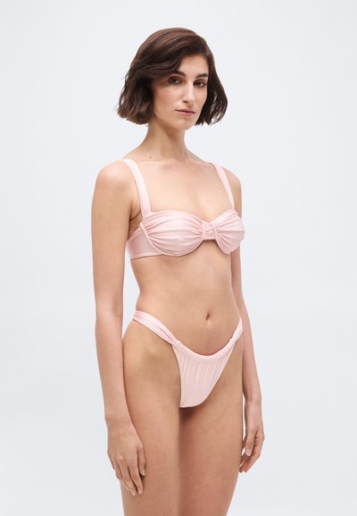 Žena s krátkymi hnedými vlasmi pózujúca v svetloružovom bikini topu s riasením a plavkových nohavičkách s vysokým strihom na bielom pozadí.