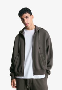 Smeđa zip-up hoodie izrađena od mekog materijala, s kapuljačom, prednjim džepovima i rebrastim manžetama. Uparena s bijelom majicom s okruglim izrezom.