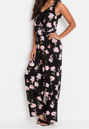 Femme portant une combinaison fleurie noire sans manches avec des fleurs roses et blanches, debout avec une main sur la hanche et des sandales noires.
