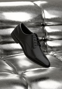 Zapato de cuero negro con punta redonda, con un diseño de cordones y costuras sutiles, posicionado sobre una superficie plateada reflectante.