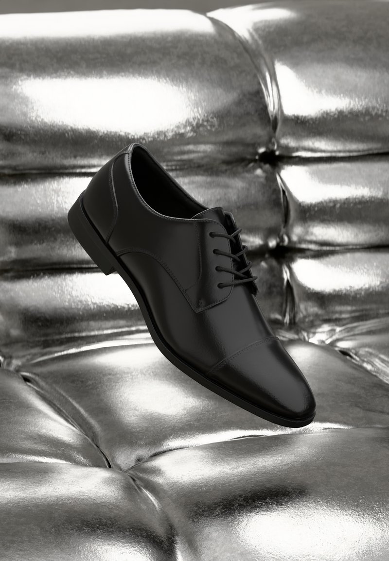Zapato de cuero negro con punta redonda, con un diseño de cordones y costuras sutiles, posicionado sobre una superficie plateada reflectante.