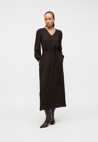 Γυναίκα με πλεξούδες, φορώντας μακρύ μαύρο φόρεμα με ζώνη και μυτερά μαύρα δερμάτινα μποτάκια, στέκεται μπροστά σε απλό φωτεινό φόντο.