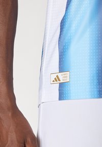 Close-up van een persoon die een blauw-wit sportshirt draagt met een gouden Adidas-label "Authentic Licensed Product" vlak bij de taille.