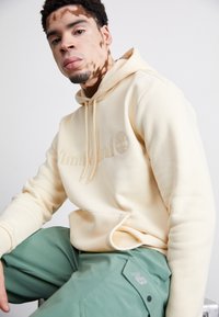 Tejfehér kapucnis pulóver, dombornyomott Timberland logóval, mentazöld cargo nadrággal párosítva, ami zsebet és New Balance logót tartalmaz. Simaság és kellemes tapintás jellemzi.