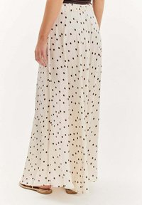 Žena oblečená v krémovej maxi sukni s hnedými bodkami, v kombinácii s plochými sandálmi, stojaca na uniformnom pozadí.