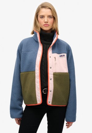 Woman wearing a blue, pink, and olive fleece jacket over a black cable knit turtleneck and black pants with a silver belt buckle.