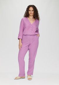 Lavendelkleurige twee-delige outfit bestaande uit een knoopsluiting, V-hals shirt met opgerolde mouwen en bijpassende, ontspannen pasvorm broeken. Gelijk gedragen met platte sandalen.