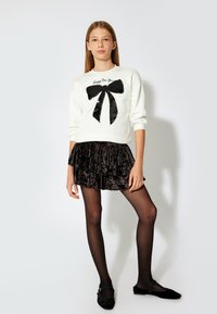 Weißer Pullover mit schwarzem Pailletten-Schleifenmotiv; geschichteter schwarzer Samtrock; schwarze Strumpfhose mit Punkten; schwarze Ballerinas mit dünnem Riemen.