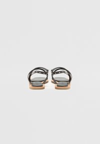 Sandales en cuir noir avec des brides croisées, des accents en clous dorés et une semelle plate en caoutchouc beige. Semelle intérieure lisse au design minimal.