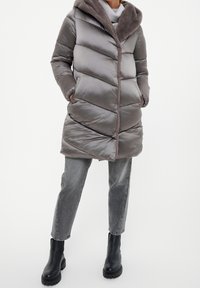 Abrigo acolchado gris con capucha forrada de piel, que presenta un patrón acolchado en forma de chevrones, bolsillos delanteros y cierre con cremallera. Se lleva con vaqueros grises y botas negras.