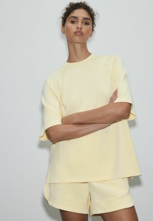 Femme aux cheveux bouclés portant un haut oversize à manches courtes jaune pâle et un short assorti, debout les bras croisés devant un fond clair.