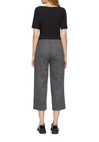 Schwarzes, kurzärmliges Oberteil und graue, gesprenkelte weit geschnittene Hose. Die Hose hat einen strukturierten Stoff; die Schuhe sind glänzend schwarz mit dickem Absatz.
