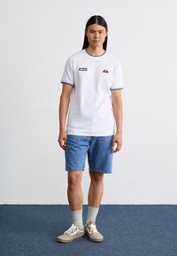 Hvid t-shirt med korte ærmer, navy trim og logo, parret med lyseblå denimshorts. Grå sokker og beige sneakers fuldender looket.