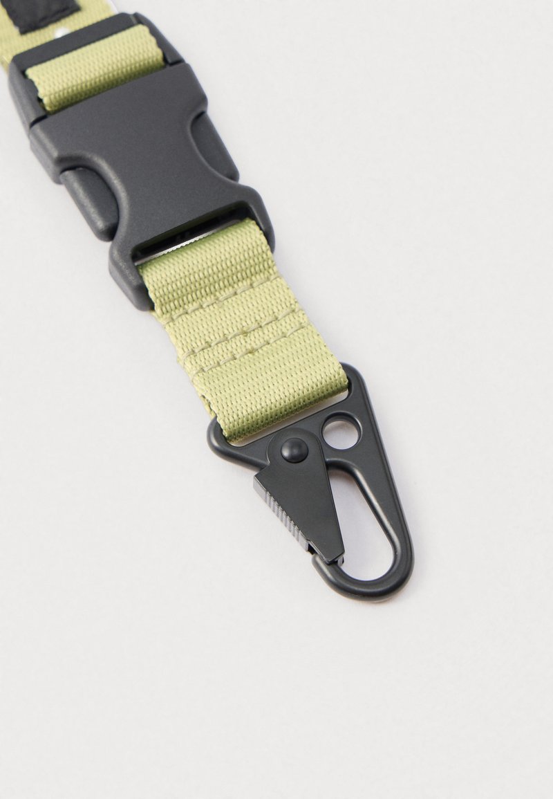 Cinturino regolabile in tessuto verde chiaro con fibbia e gancio in plastica nera. Presenta una superficie texturizzata e un design robusto e resistente.