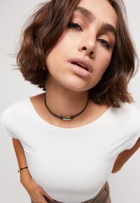 Collier ras du cou en cuir noir avec un pendentif rectangulaire en turquoise, maintenu par de petites perles marron. Porté sur une chemise blanche.