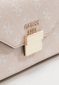 Beżowa torebka teksturowana z jasnym białym wzorem, złoty logo "GUESS 1981" oraz prostokątny złoty zamek na przedniej klapie.