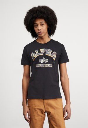 T-shirt en coton noir avec "ALPHA INDUSTRIES" imprimé en texte multicolore ; col rond ; manches courtes ; porté avec un pantalon beige.