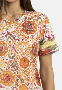Blusa corta de manga corta colorida con un vibrante patrón floral y geométrico en naranja, rosa, verde y amarillo sobre un fondo blanco. Diseño de escote en V.