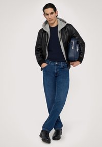 Svart läderbombarjacka över en grå hoodie, marinblå tröja, blå jeans och svarta stövlar. Håller en blå väska med silverdetaljer.